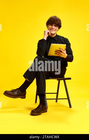 Young attractive cheerful man, businessman sitting on chair holds tablet and looking at camera against vivid yellow studio background. Stock Photo