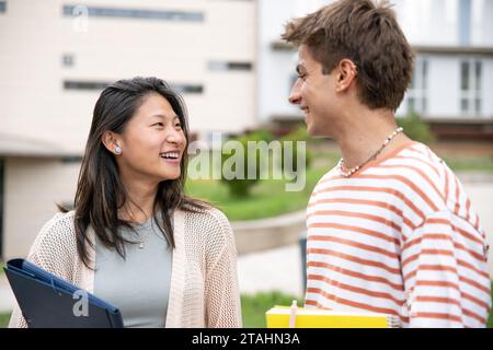 Diverse couple smiling and holding folders in a campus.Two young students looking each other smiling and satisfied in the street. Stock Photo