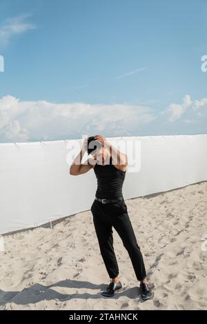 cuban man in black outfit touching hair while standing on sand on beach in Miami Stock Photo