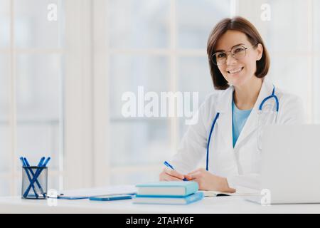 Smiling female doctor at desk with laptop, taking notes in bright office. Stock Photo