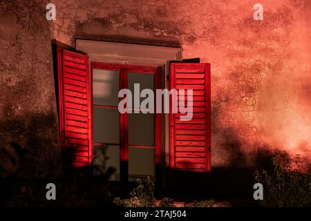 A pair of bright red shutters, attached to a window on an old farmhouse in Tuscany, Italy,  are illuminated by artificial light. Stock Photo