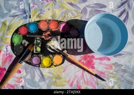 Watercolor paints, brushes and artist's palette on a flowered tablecloth with fresh bowl of water. Stock Photo