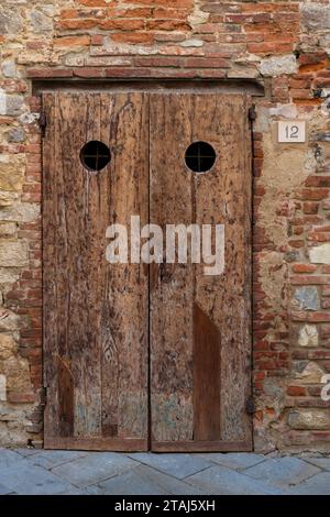 An old, wooden, unpainted, textured door with two round openings at the base of a deteriorating, red-brick building in the village of Lucignano, Tusca Stock Photo