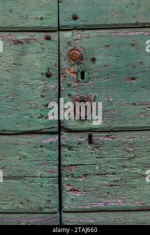 A faded, peeling and weathered, light green painted door with chipped surface, keyholes, lock and metal rusted knocker. Stock Photo