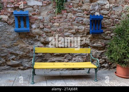 A yellow wooden bench set between two wood palettes painted blue and mounted to an aging stone and brick wall in Sinalunga, Tuscany, Italy. Stock Photo