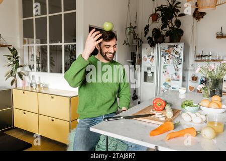 Cheerful and bearded man in green jumper balancing with apple on head in modern kitchen Stock Photo