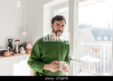 Portrait of bearded man in jumper holding cup of coffee and looking at camera in modern kitchen Stock Photo