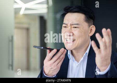 Close-up photo of an angry and serious Asian male businessman talking emotionally on the phone through a loudspeaker in the office. Stock Photo