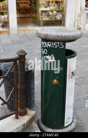 Signs of Overtourism overrunning Venice and good manners – Tourists Go Home, graffiti painted on a green rubbish bin by a popular bridge and shops Stock Photo