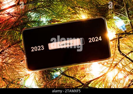 the concept of changing the year from 2023 to 2024. smartphone on the decorated fir branches in bright lights of garlands with the process of loading Stock Photo