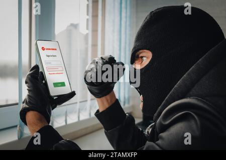 Angry young man in balaclava using cell phone. Theft of personal data. Hacking the city. A bandit in the office picks up a password to log in to a sma Stock Photo