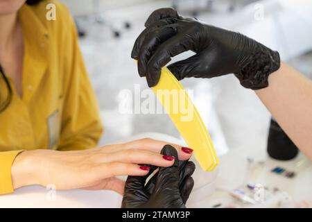 Woman in nail salon receiving manicure by beautician. Manicure process in beauty salon, close up. Manicurist filing client's nails at table, closeup. Stock Photo