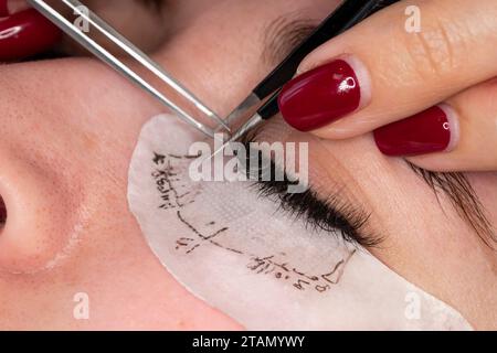 Eyelash extension procedure. Master tweezers sets fake lashes on beautiful woman close-up. Stock Photo