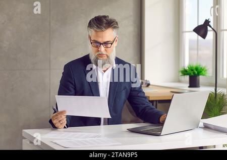 Mature businessman working with paper document and using laptop in his office Stock Photo