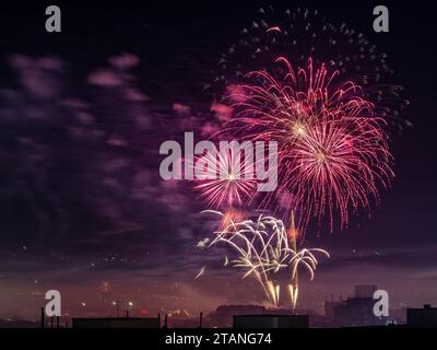 Red fireworks burst in the midnight sky. Festive fireworks explosions over small city celebrating holiday event. Pyrotechnic and firecracker concept. Stock Photo