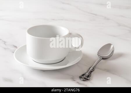 White cup on saucer and teaspoon over marble table. Empty clean porcelain crockery for drink design mockup. Tableware, breakfast, tea coffee concept. Stock Photo