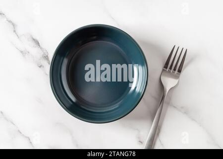 Dark blue ramekin and fork over marble tabletop. Empty clean ceramic bowl for food design mockup. Modern porcelain crockery and tableware concept. Stock Photo