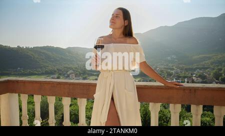 Young woman in a flowing dress relishes a glass of red wine on the balcony or villa terrace, while taking in the magic of a mountain sunset. Travel, s Stock Photo