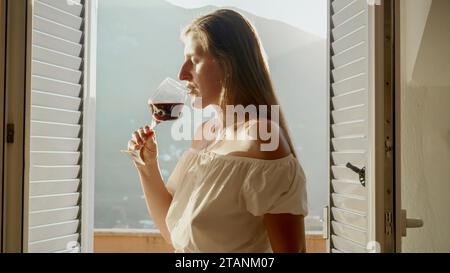 Lady stands on the villa terrace, sipping red wine from a glass, while the scenic mountains serve as a captivating backdrop. Stock Photo