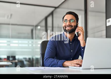 Happy busy Indian businessman talking on cell phone using laptop in office. Stock Photo