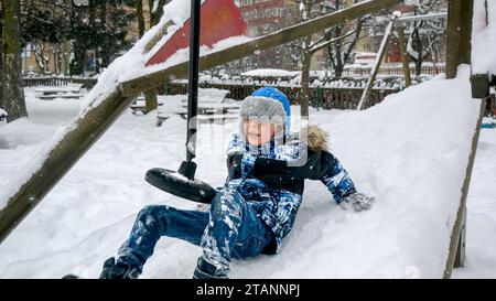 Little boy falling on the playground covered with snow from the zipline. Children playing and having fun outdoors during winter holidays and weekend. Stock Photo