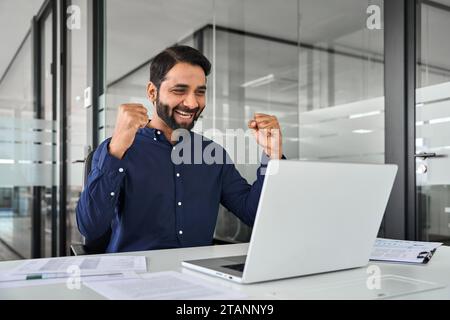Happy Indian business man winning prize online working using laptop in office. Stock Photo