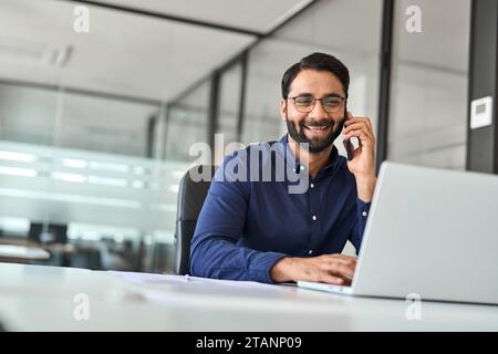 Happy Indian businessman merchant calling on cell phone using laptop in office. Stock Photo