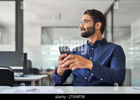 Happy professional Indian businessman employee looking aside holding smartphone. Stock Photo