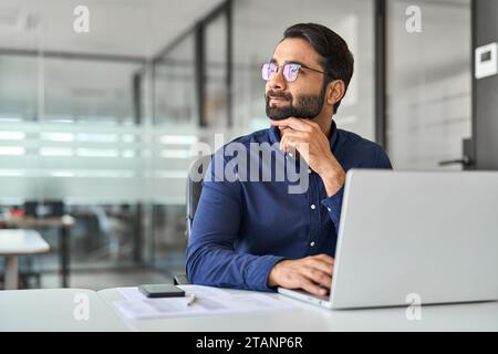 Happy Indian business man worker thinking looking aside working on laptop. Stock Photo