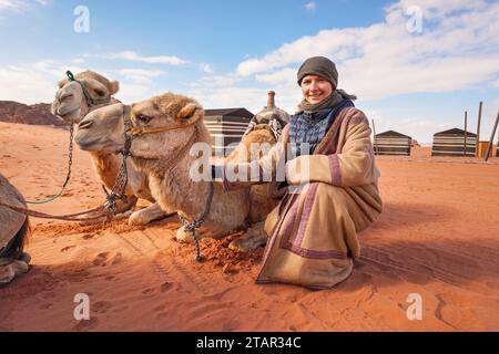 Young woman in traditional Bedouin coat - bisht - and headscarf crouching next to two camels laying on red desert ground, smiling, camp tents in backg Stock Photo