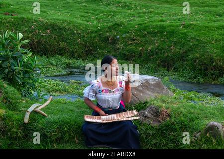indigenous girl sitting next to a river tasting a large guaba, the girl wears a traditional dress from her culture. Stock Photo