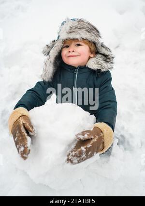 Snowman and funny child the friend is standing in winter hat and scarf with red nose. Joyful kid Having Fun with snowman in Winter Park. Winter Stock Photo