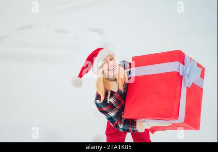 Winter delivery service. Woman carrying a big present isolated on snow winter background. Portrait of Santa woman with huge red gift looking at camera Stock Photo