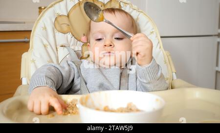 Portrait of little baby boy getting dirty and messy while eating porridge with spoon. Stock Photo