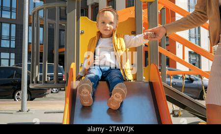 Cute boy holding mothers hand and riding down the slide on playground. Happy parenting, family having time together, kids and parents outdoors Stock Photo