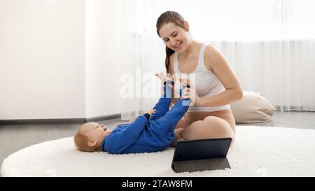 Young caring mother doing stretching and fitness exercises to her baby son lying on floor at living room. Stock Photo