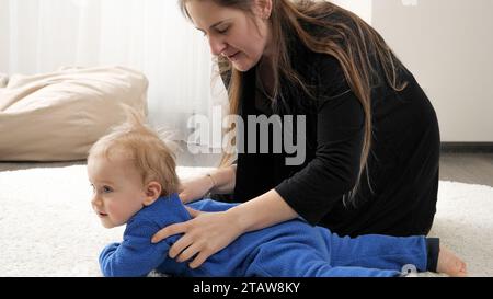 Young caring mother doing gymnastics and stretching exercises to her little baby boy on floor in living room. Family healthcare, active lifestyle, par Stock Photo