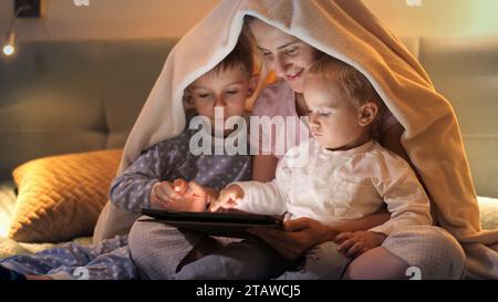 Happy family sitting under blanket and using tablet computer at night. Family having time together, parenting, happy childhood and entertainment Stock Photo