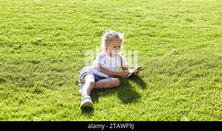 Little girl sitting on the lawn in the summer city park and holding smartphone. Childhood, leisure and people concept - happy child rest and have a go Stock Photo