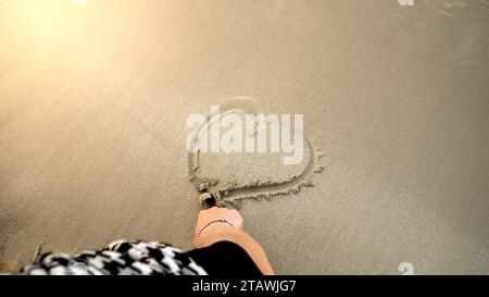 Closeup of female hand drawing a heart shape of wet sand at sea beach. Concept of loving travel, tips, journey and tourism Stock Photo