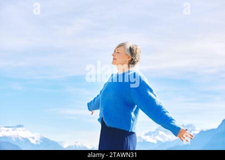 Outdoor portrait of happy middle age 55 - 60 year old woman with arms open wide, posing on blue sky and mountain background Stock Photo