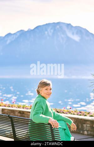 Portrait of middle age woman enjoying nice day outside, relaxing on bench in front of mountain lake Stock Photo