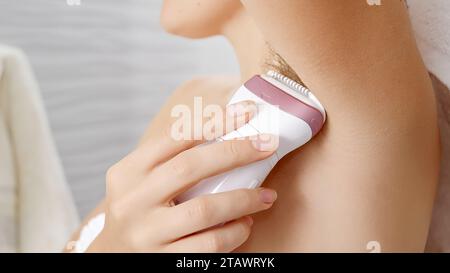 Young woman using electric epilator to remove armpit hair in bathroom. Stock Photo
