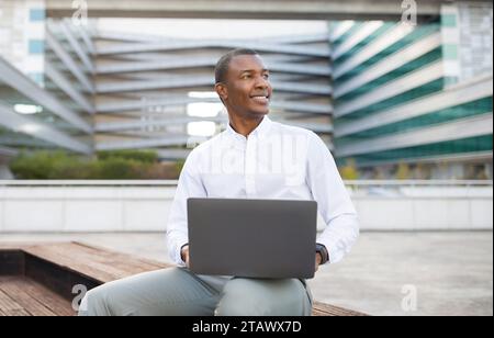 Smiling black businessman using laptop while sitting on outdoor bench Stock Photo