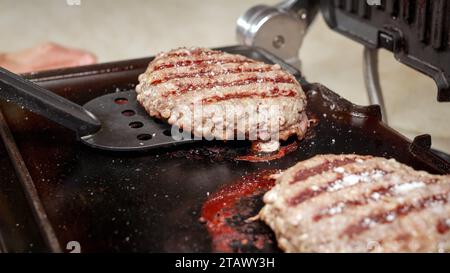 Closeup of turning over beef burger patties cooking on electric grill. Cooking at home, kitchen appliance, healthy nutrition, hamburger ingredients Stock Photo