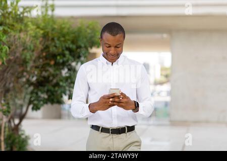 Portrait Of Smiling Young Black Businessman In Shirt Using Smartphone Outdoors Stock Photo