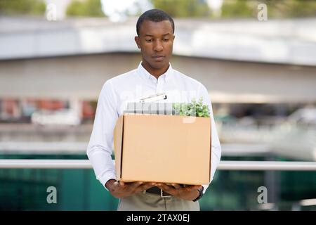 Depressed black man in shirt walking with cardboard box of belongings outdoors Stock Photo