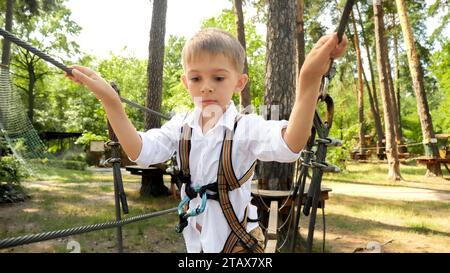 Little boy with safety rope walking over the wooden bridge strung in adventure park. Stock Photo