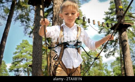 Portrait of little boy smiling while walking over strung rope in climbing training center. Active childhood, healthy lifestyle, kids playing outdoors, Stock Photo