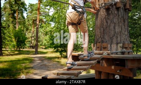 Child's feet walking over wooden rope bridge strung high between trees in forest. Stock Photo